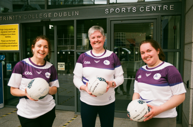 Three females in sports attire, each holding a Gaelic football, standing in front of the entrance to the Trinity College Dublin Sports Centre.