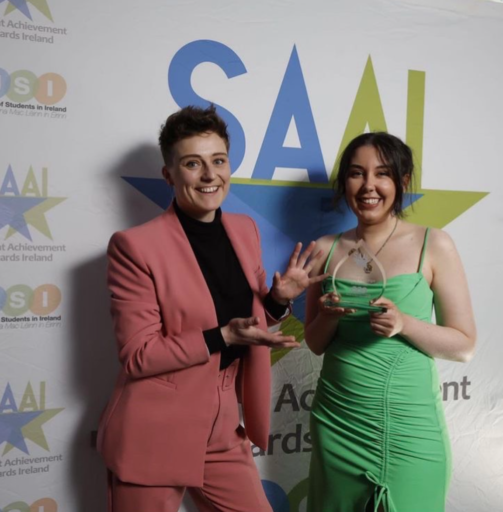 Rachel, a white woman with dark brown hair accepting the Equality Campaign of the Year Award. She is wearing a green dress, standing next to a person in a pink suit. 