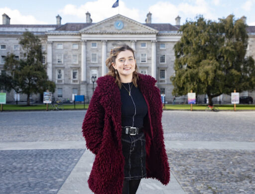 Courtney, a white woman with tied up light brown hair standing in front square of Trinity College Dublin